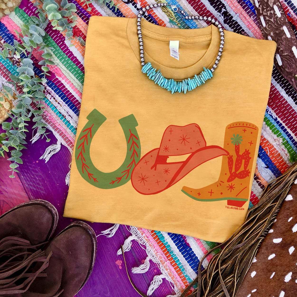 This mustard yellow graphic tee features three hand handdrawn cowgirl necessities - a horseshoe, a cowboy hat, and a boot in various pops of colors. This is a Bella + Canvas tee with short sleeves and a crew neckline. It is shown here styled as a flat lay with a turquoise necklace and brown booties.