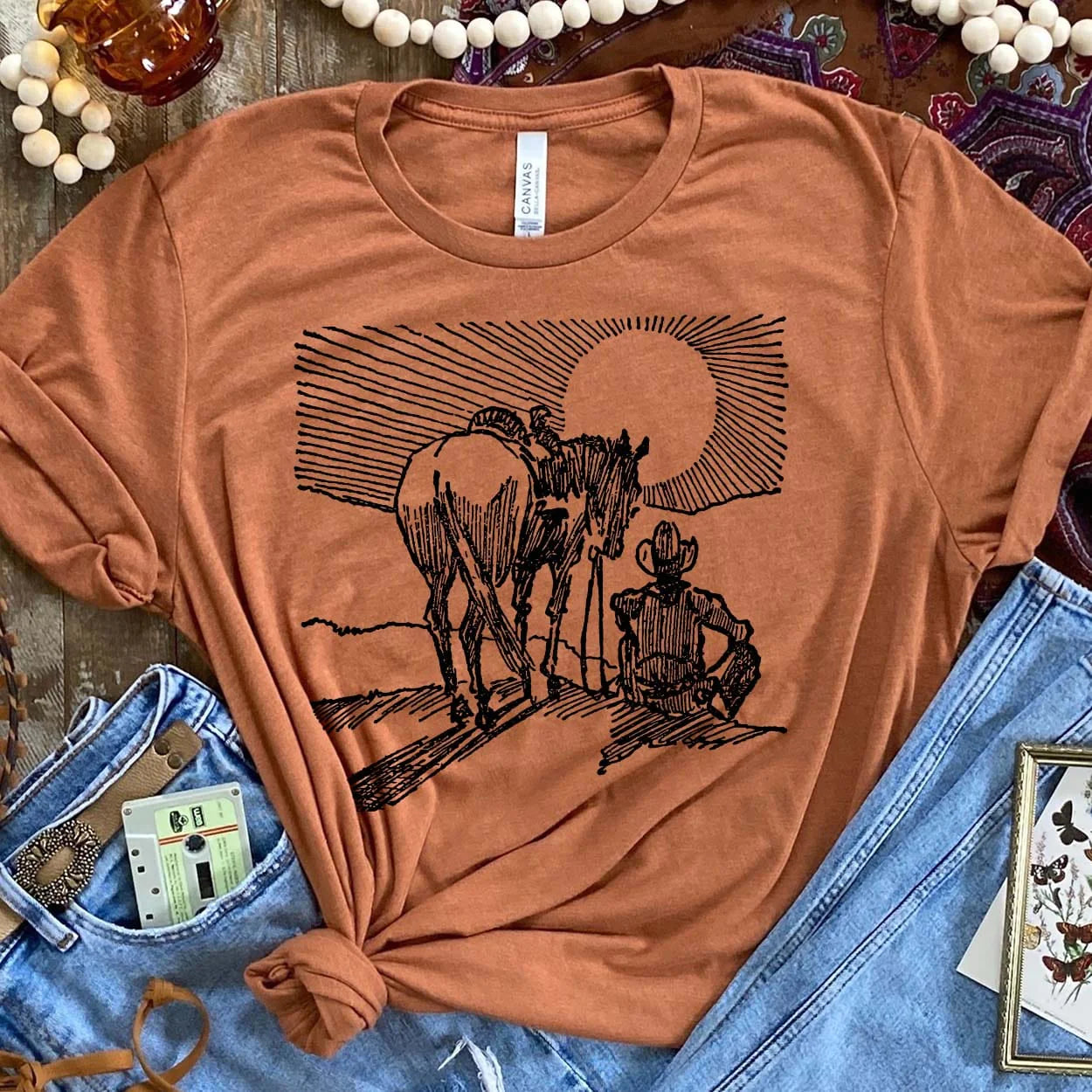 A light brown short sleeved shirt with a centered black and white graphic of a cowboy and a horse watching the sunset. Item is pictured on a brown background.
