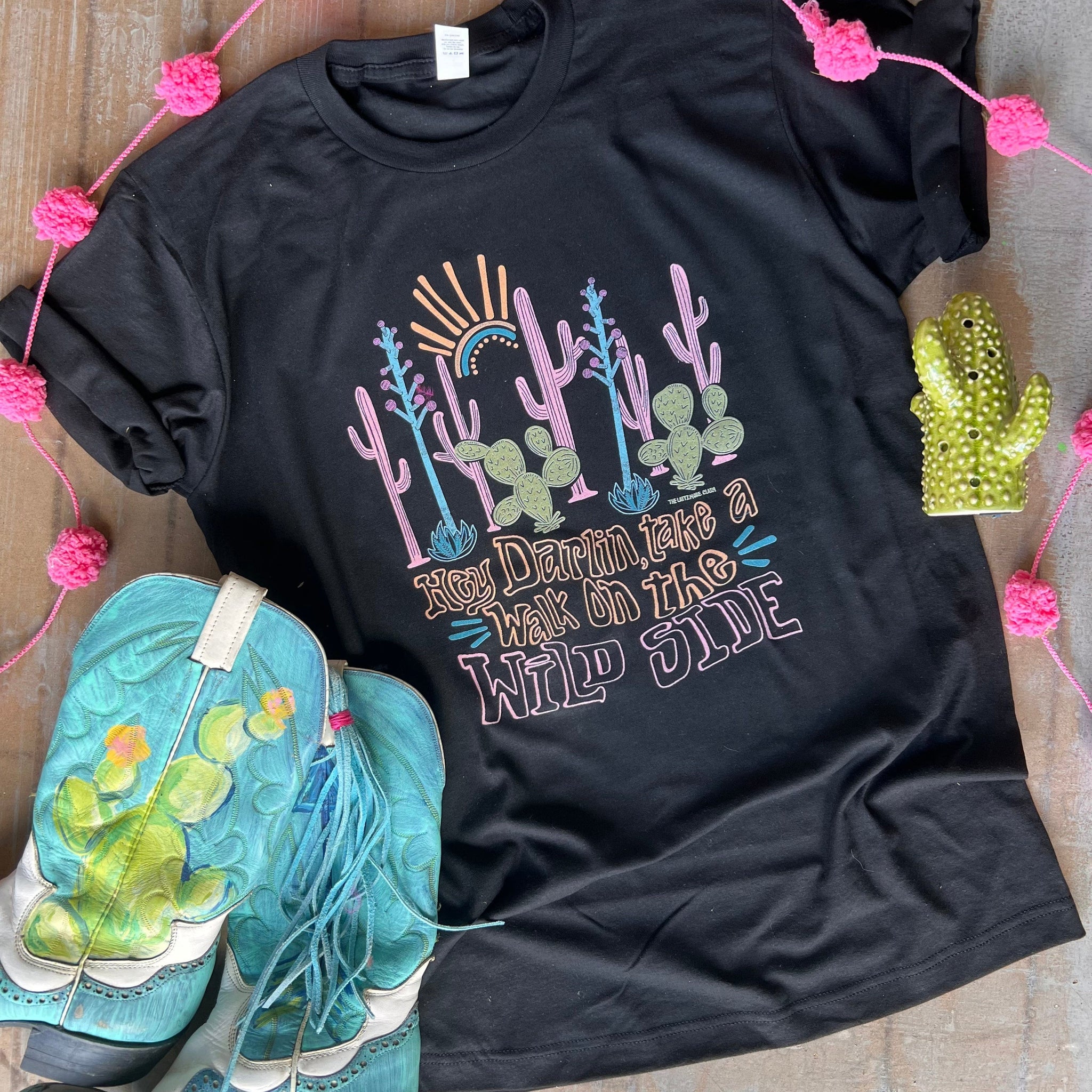 This Black Bella + Canvas tee includes a crew neckline, short sleeves, and a hand drawn graphic with words "hey darlin, take a walk on the wild side" in orange and pink. The graphic is a variety of colorful cacti and a partial sun. 