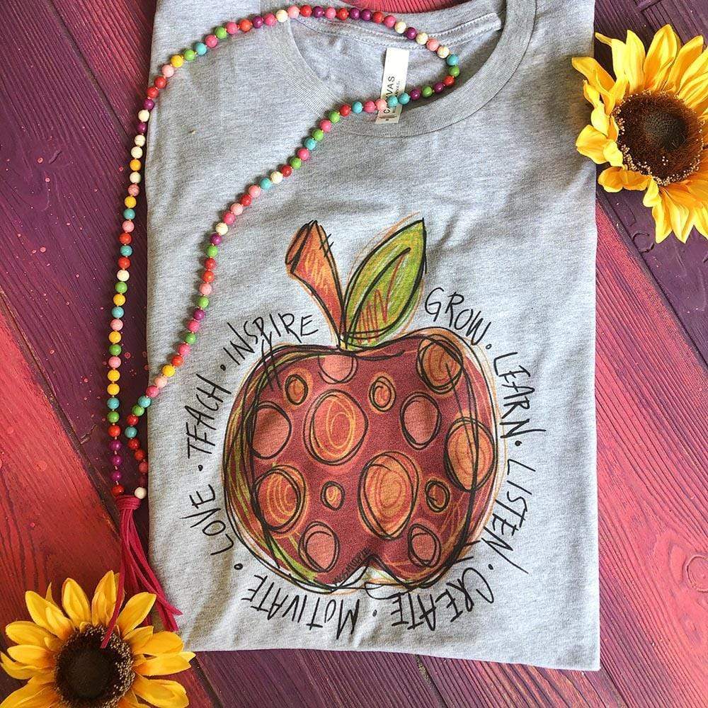 This Grey Bella + Canvas tee includes a crew neckline, short sleeves, and a hand drawn red and orange polka dot apple with inspirational words in black surrounding the apple. Words include "grow, learn, listen, create, motivate, love, teach, inspire". 