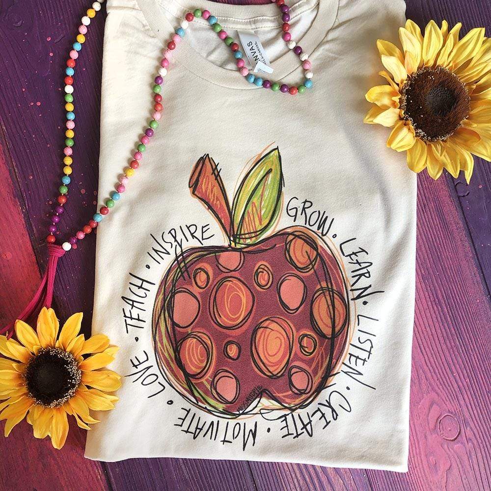 This Cream Bella + Canvas tee includes a crew neckline, short sleeves, and a hand drawn red and orange polka dot apple with inspirational words in black surrounding the apple. Words include "grow, learn, listen, create, motivate, love, teach, inspire". 