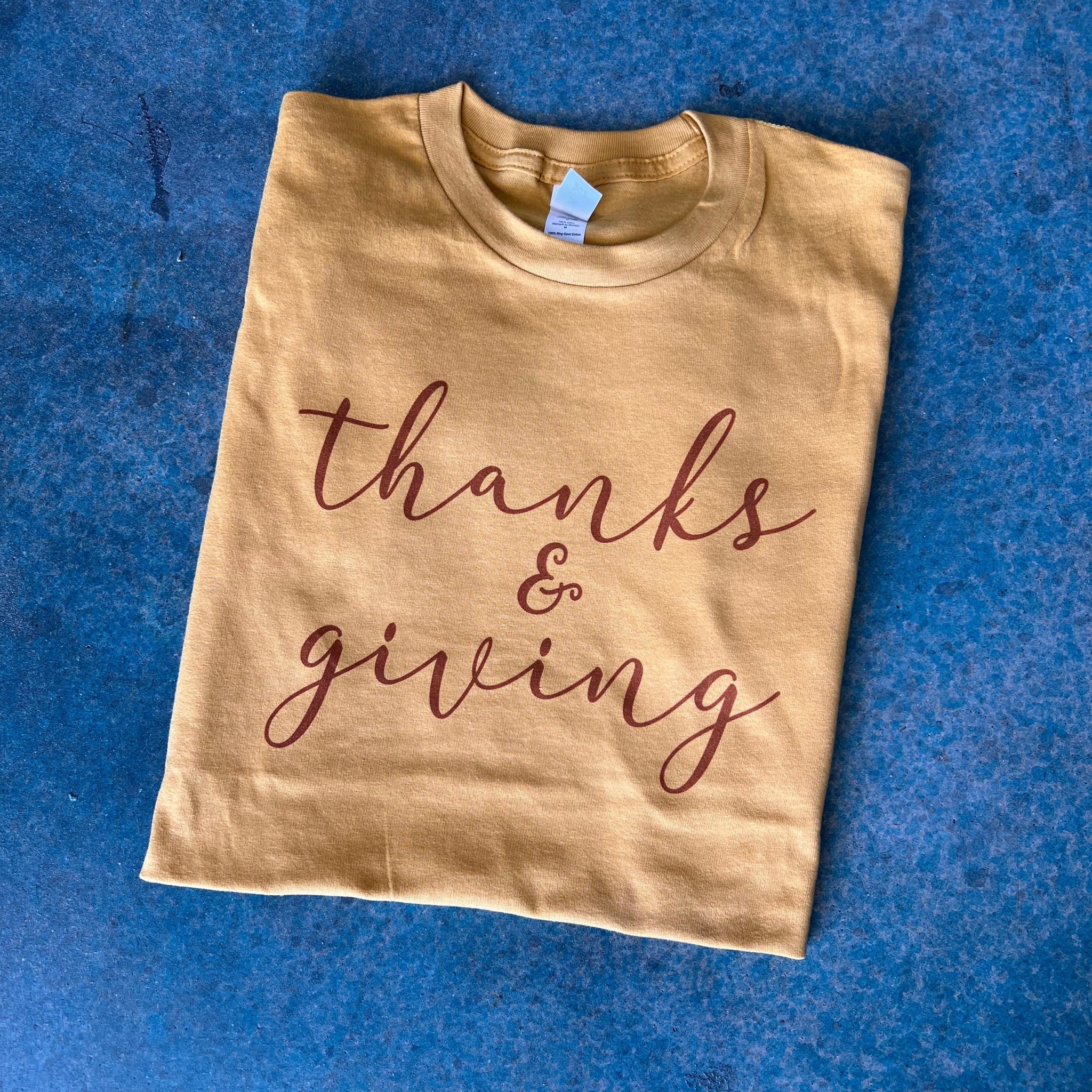 This mustard yellow tee includes a crew neckline, short sleeves, and a handwritten, rust red, cursive font that says "thanks & giving". It is pictured here as a folded flatlay on a blue background. 