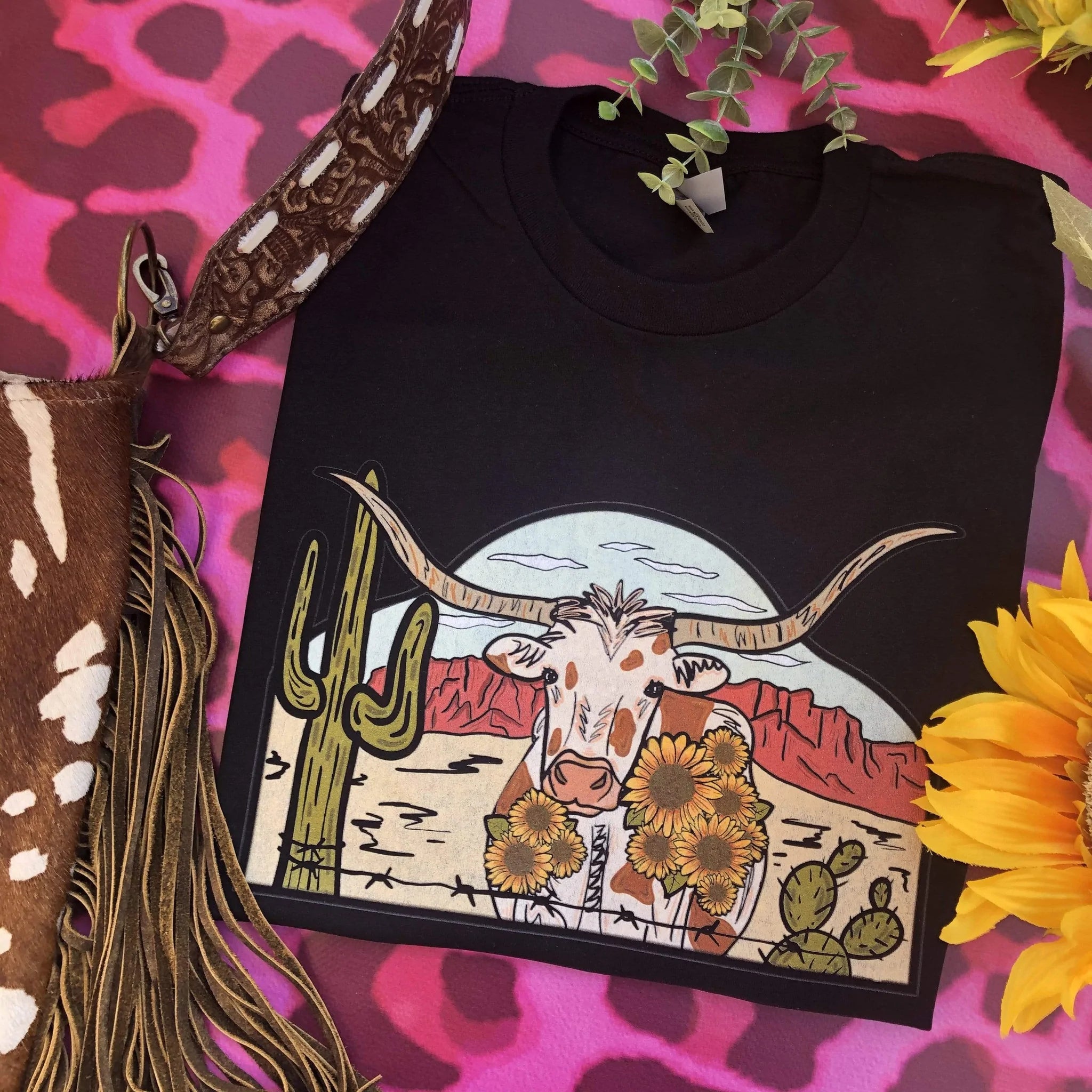 This black Bella + Canvas graphic tee includes a crew neckline, short sleeves, and cute hand drawn design of a desert scene with a longhorn in the middle. This tee is shown in this photo to be styled as a folded flat lay with a brown cow print purse. 