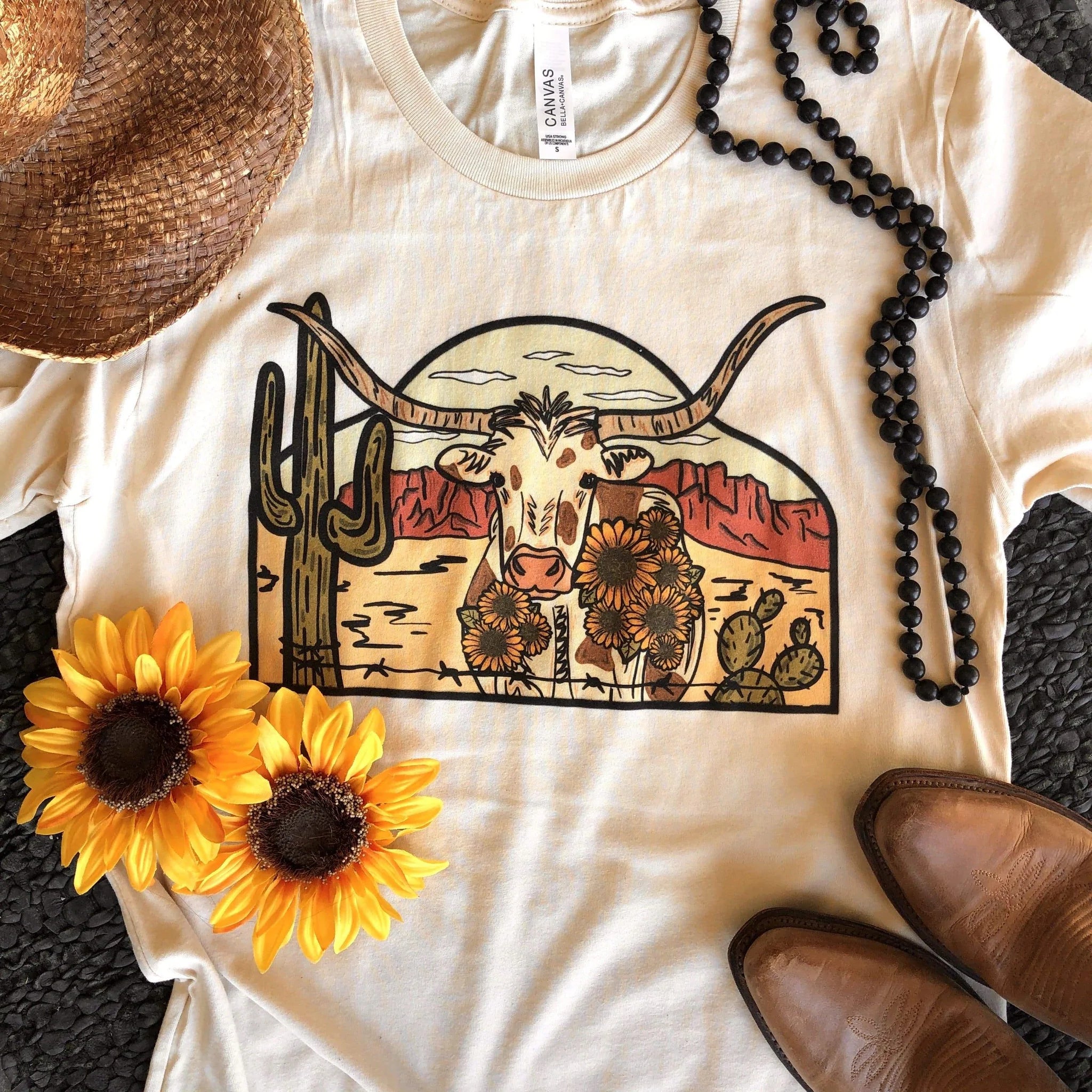 This cream Bella + Canvas graphic tee includes a crew neckline, short sleeves, and cute hand drawn design of a desert scene with a longhorn in the middle. This tee is shown in this photo to be styled as a flat lay with a light brown straw hat, black beaded necklace, and light brown boots. 