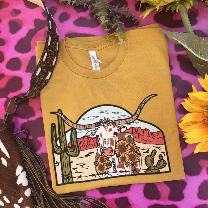 This mustard yellow Bella + Canvas graphic tee includes a crew neckline, short sleeves, and cute hand drawn design of a desert scene with a longhorn in the middle. This tee is shown in this photo to be styled as a folded flat lay with a brown cow print purse.