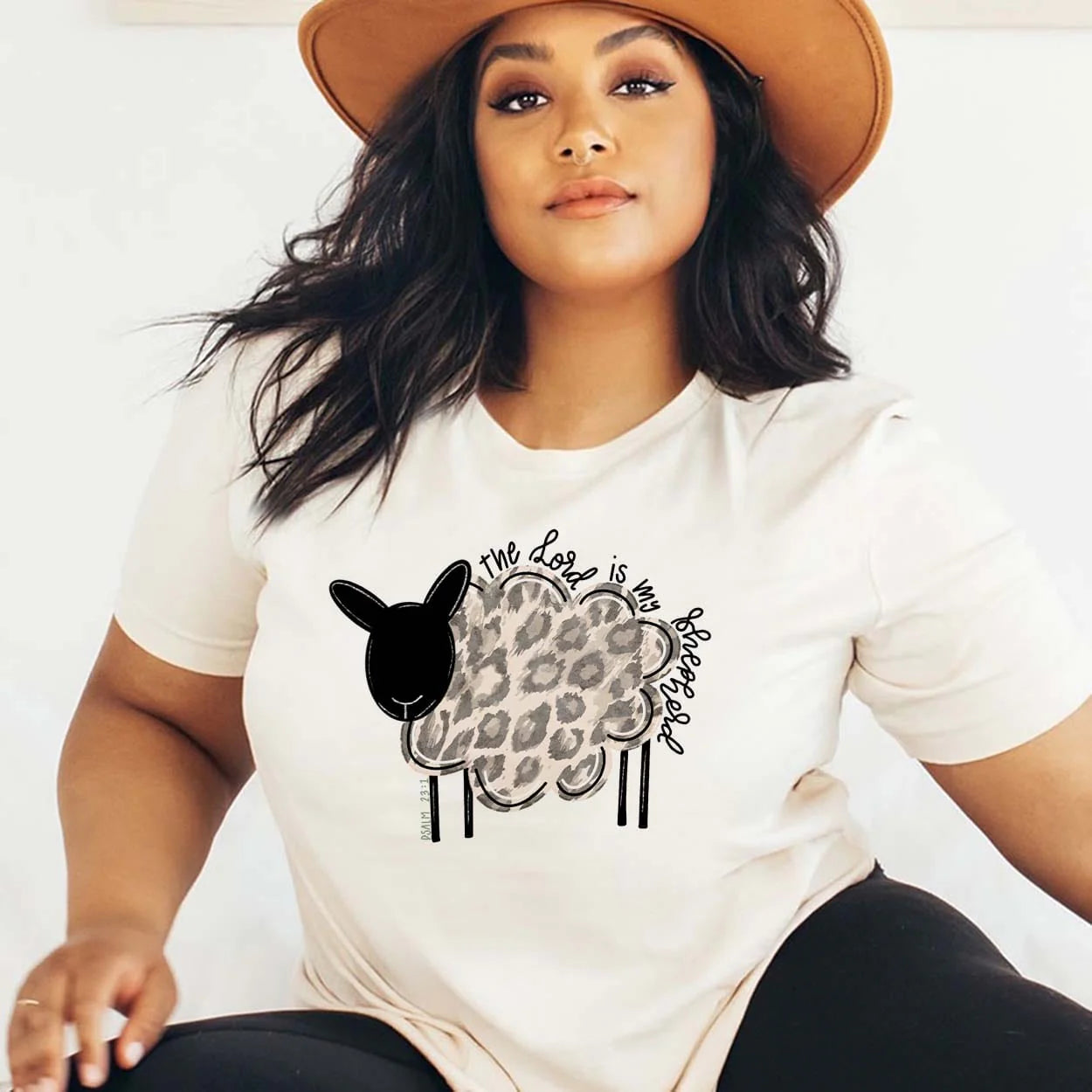 A cream short sleeve crew neck tee with a picture of a cartoon sheep, leopard print wool, and the words "the lord is my shepard" in cursive above the sheep's wool. Item is pictured on a plain white background