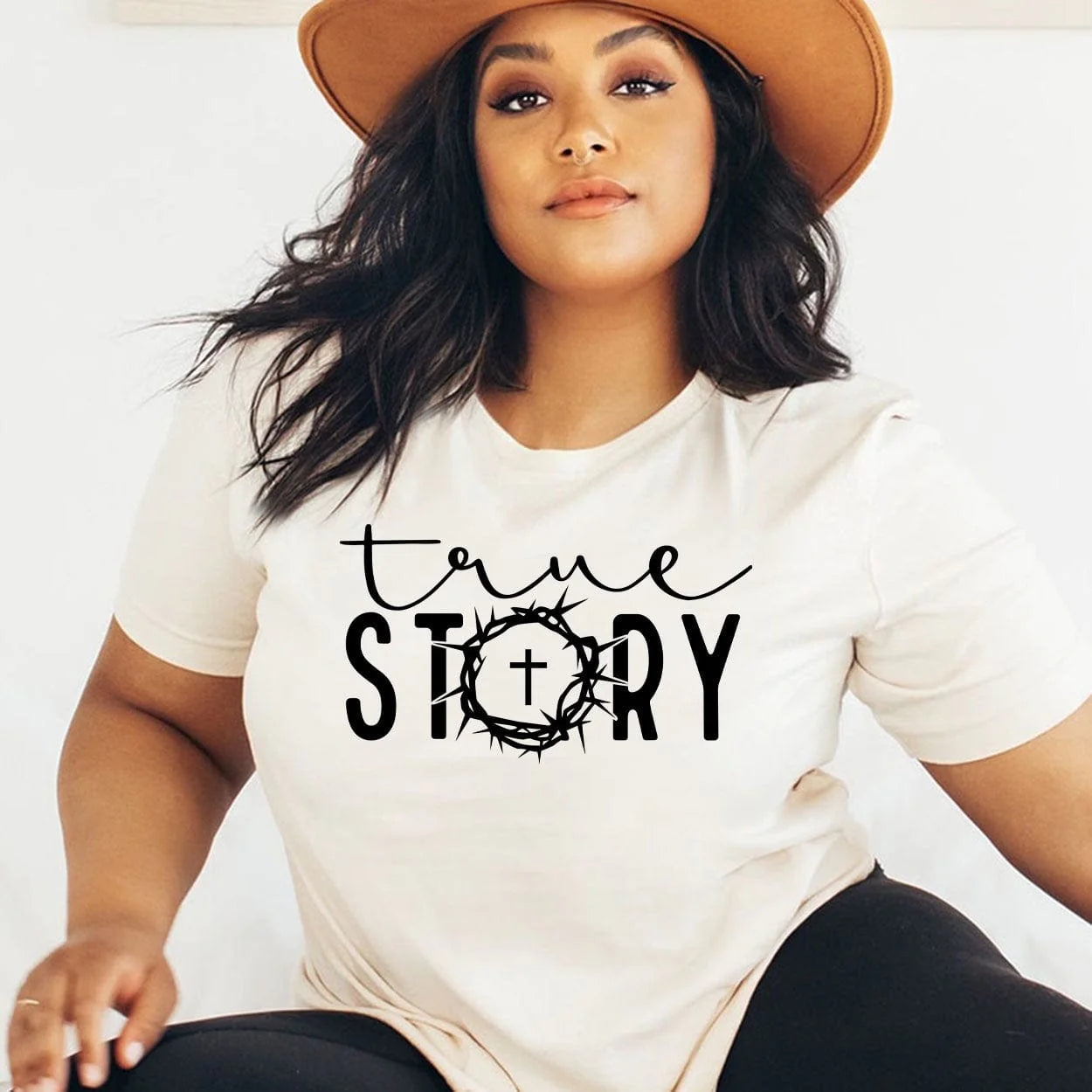 A cream colored short sleeve tee with the words "True story" in the middle. "True" being in cursive and "story" in a bold font with the O replaced with barbed wire and a cross in the center. Item is pictured on a plain white background.