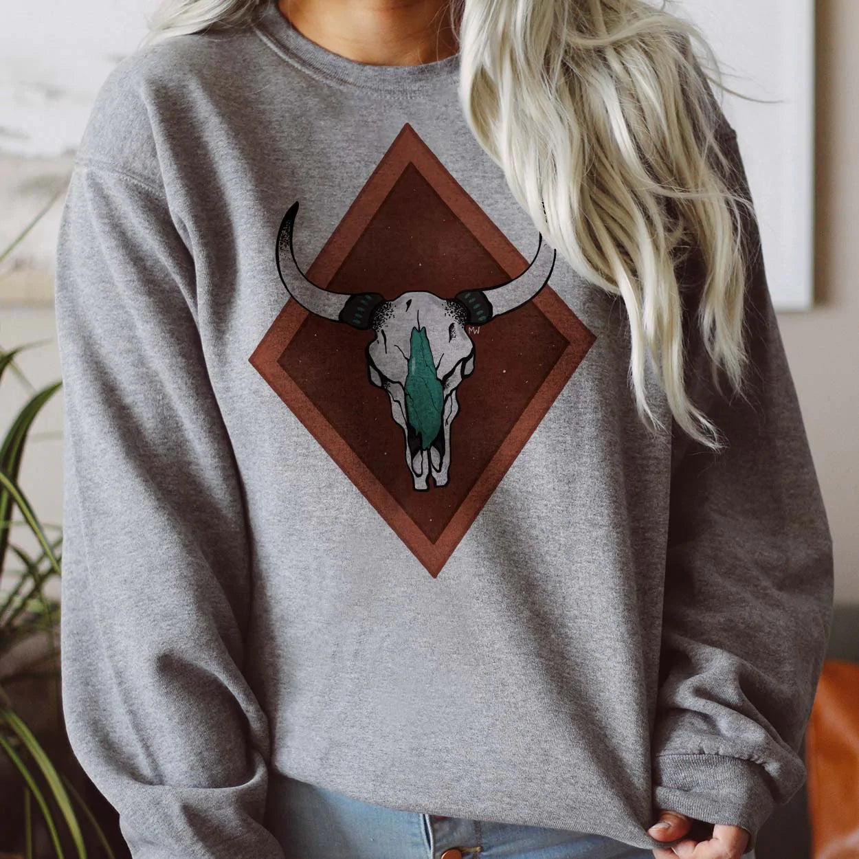 This grey sweatshirt includes a crew neckline, long sleeves, and cute hand drawn design of a detailed cow skull in white and turquoise with a two-tone rust red double diamond background. 