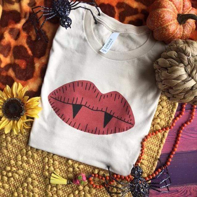 This is a white t-shirt with red lips with two vampire teeth. In the background there are two glittered spiders, two decorative pumpkins, sunflower and a beaded orange necklace with a pink skull attached.
