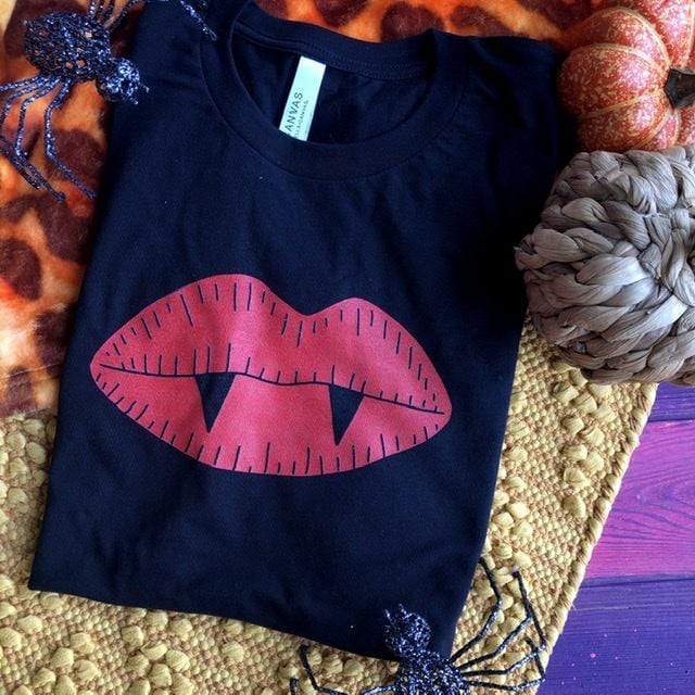 This is a black t-shirt with red lips with two vampire teeth. In the background there are two glittered spiders and two decorative pumpkins.