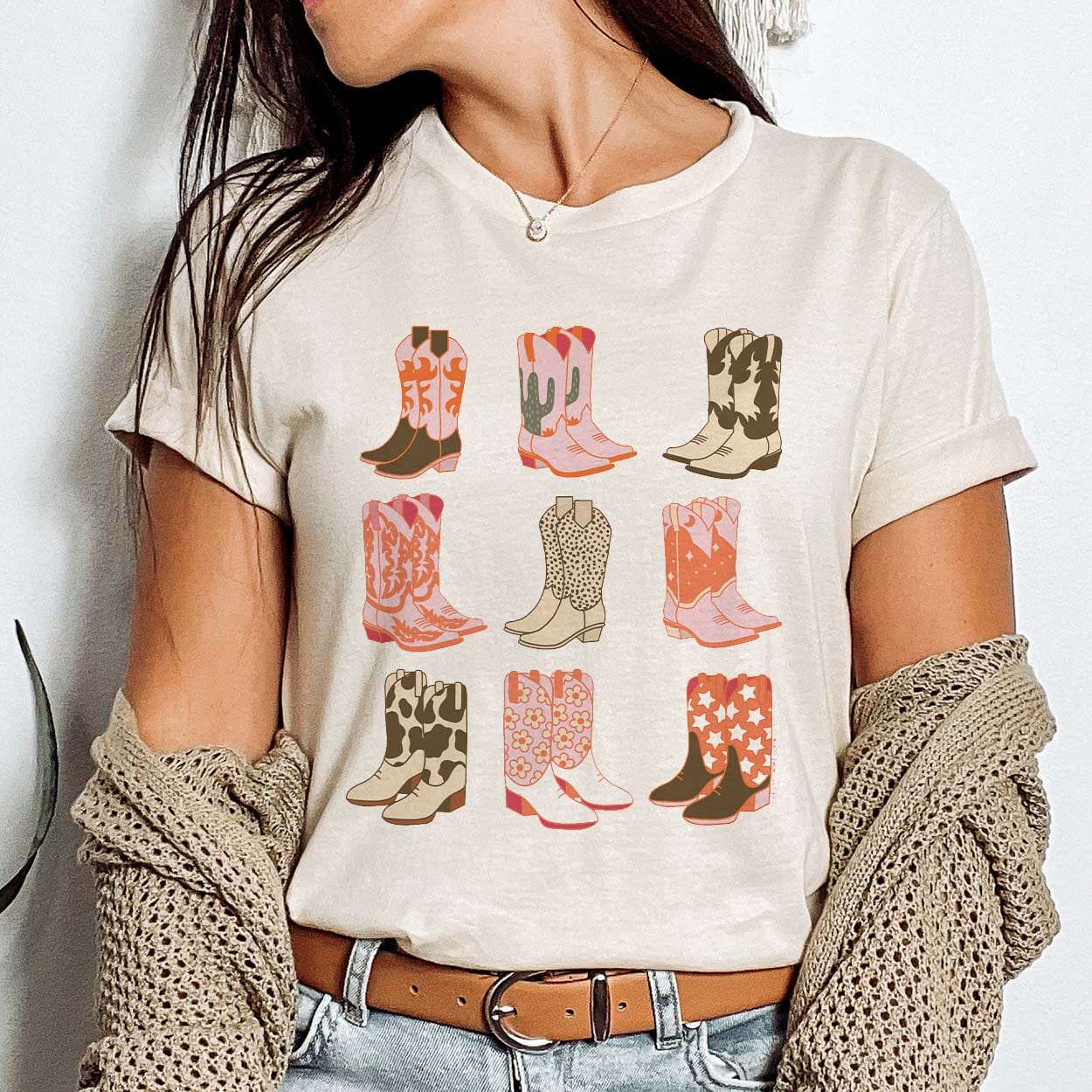 A cream colored short sleeve shirt with cuffed sleeves featuring a graphic of six different pairs of boots. The colors pink, brown, nude, orange, green, and cream are featured on the boots with orange designs, cacti, cow print, stars, polka dots, and flowers. Item is pictured on a plain white background