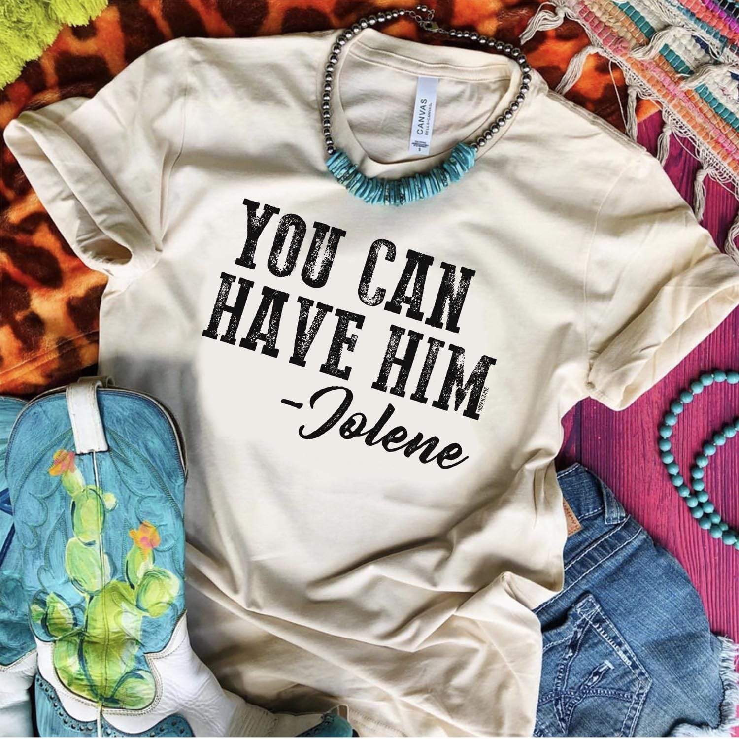 A cream colored graphic tee laid on top of different rugs. Pictured with cowgirl boots, denim shorts, and turquoise necklace. Tee shirt has a graphic that says "You can Have Him" - Jolene.